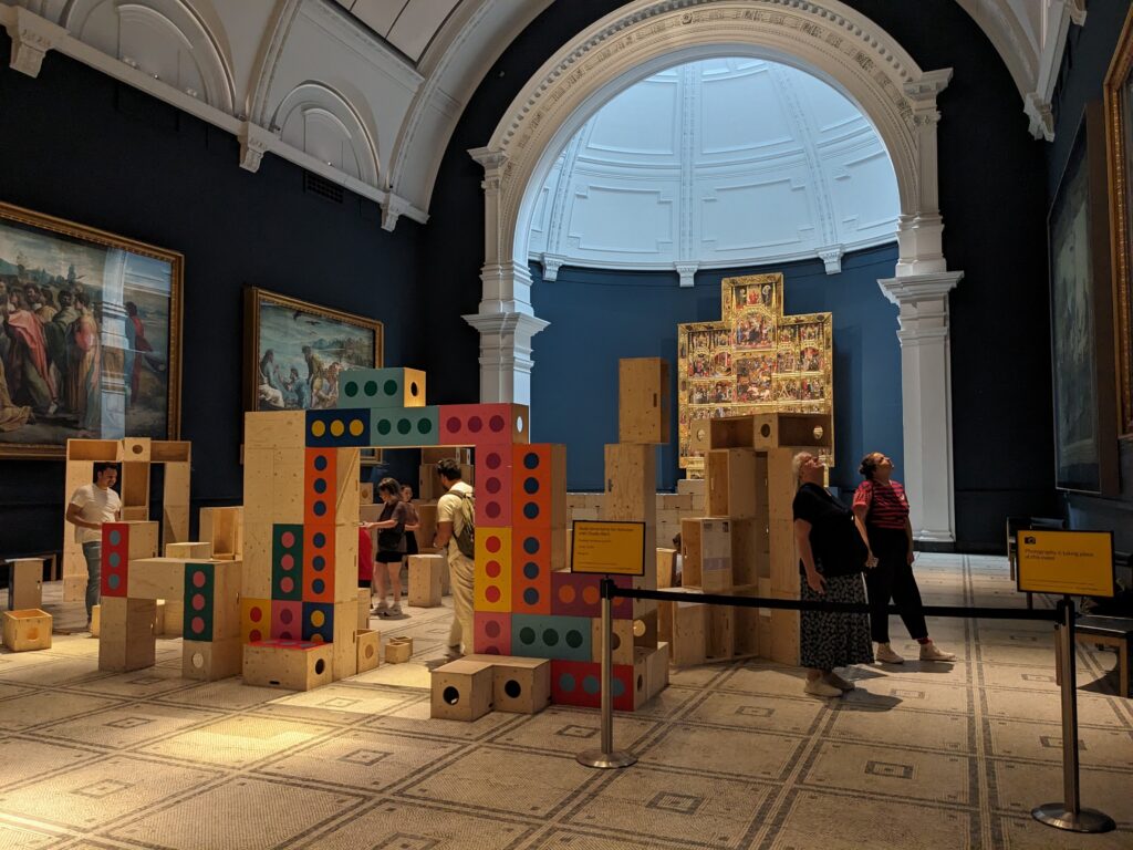 People observe a colourful wooden block installation in a museum gallery with large paintings and an ornate altarpiece. High arched ceilings and detailed architecture frame the spacious room, reflecting the spirit of Upstart within the renowned halls of the V&A.