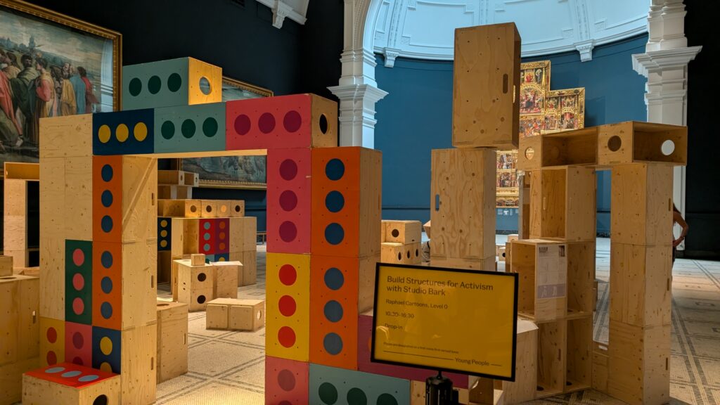 Large wooden blocks with colourful dot patterns are assembled into towering structures in a V&A museum gallery, surrounded by classic paintings and a sign inviting visitors to Build Structures for Activism with 3D modular blocks, inspired by the Upstart initiative.