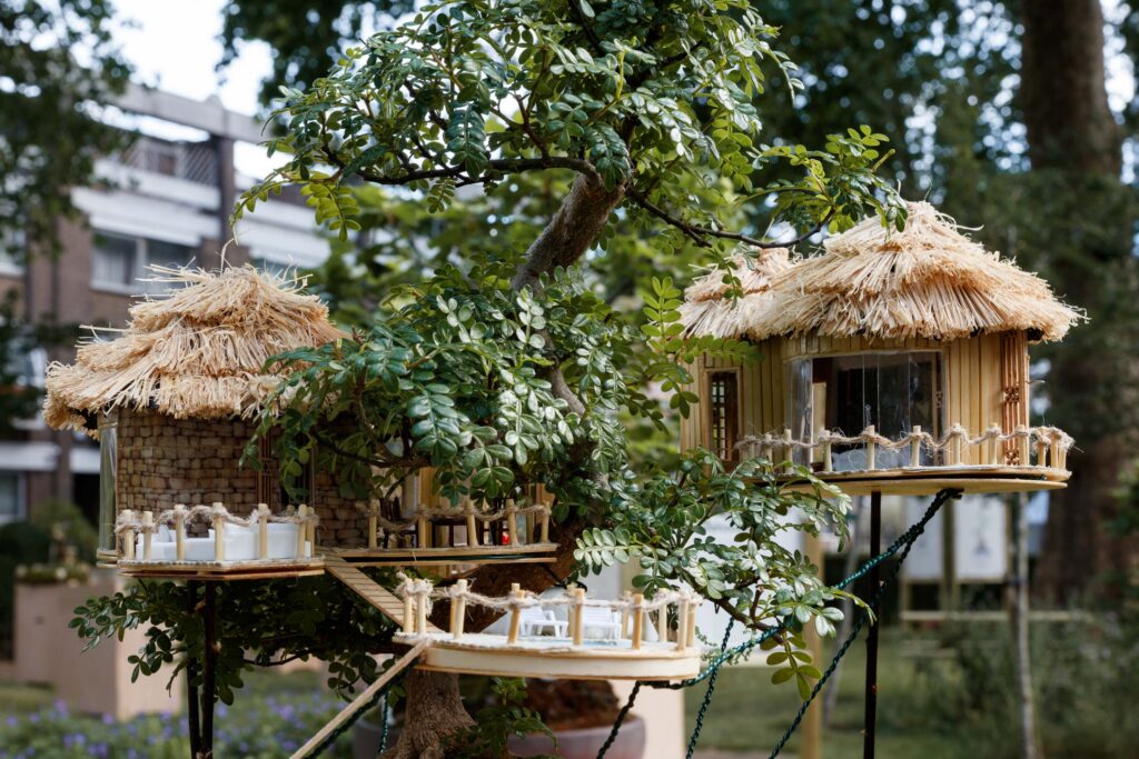 Two miniature treehouses with straw roofs and tiny furniture sit nestled in the branches of a lush bonsai tree, surrounded by vibrant greenery. A residential building is blurred in the background, evoking the artful serenity found at a bonsai exhibition.