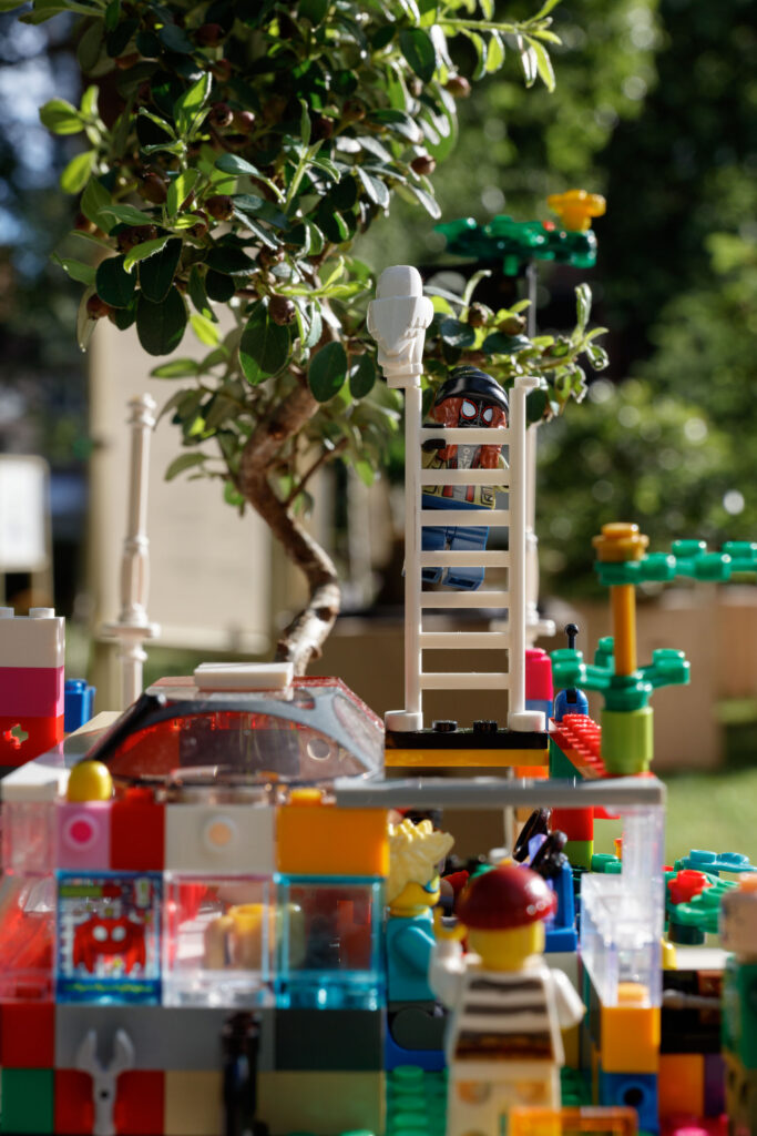 Lego figures and colourful bricks form a playful scene outdoors. One figure climbs a white ladder towards a small bonsai tree, while others stand below amid greenery and bright sunlight.