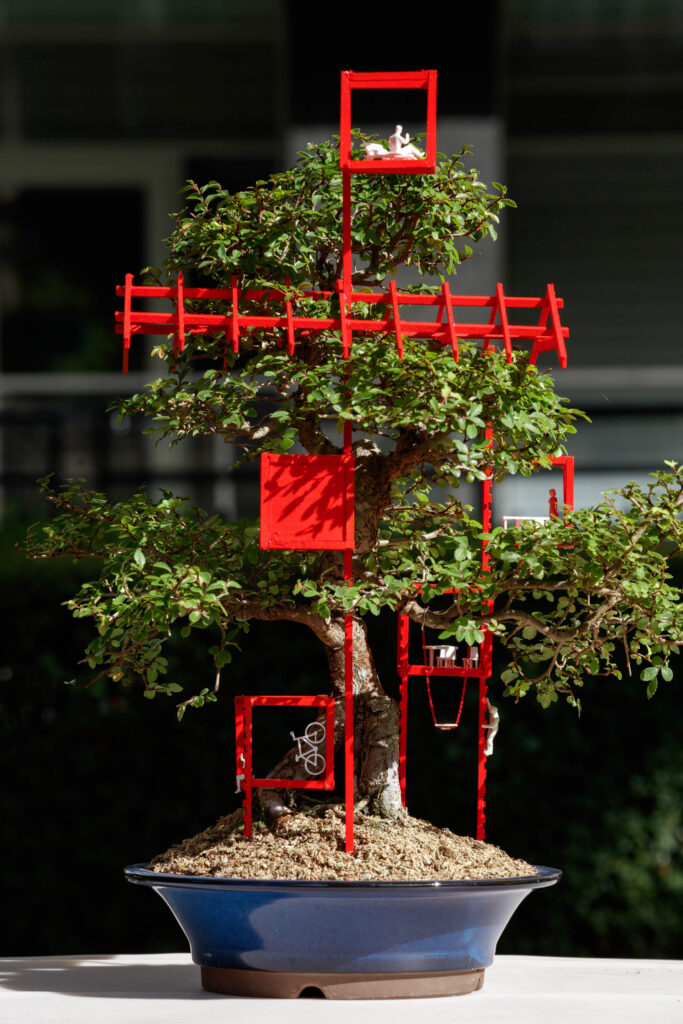 A bonsai tree in a blue pot features red geometric structures and tiny white figurines arranged among its branches, creating an eye-catching display perfect for a bonsai exhibition.