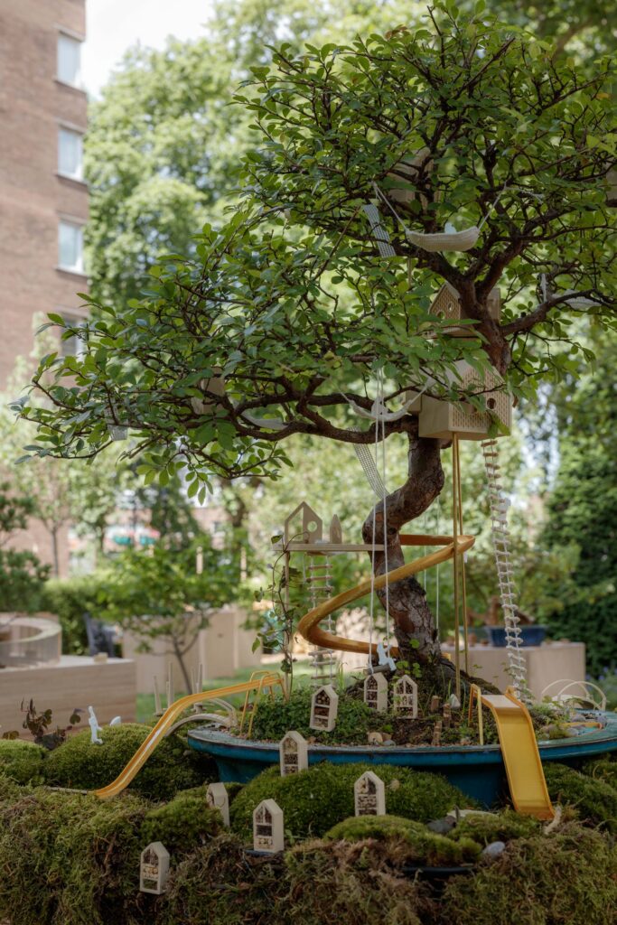 A bonsai tree decorated with miniature fairy houses, a yellow slide, bird boxes, and a small ladder creates a whimsical outdoor scene at a Bonsai Exhibition, set against lush greenery and nearby buildings in the background.