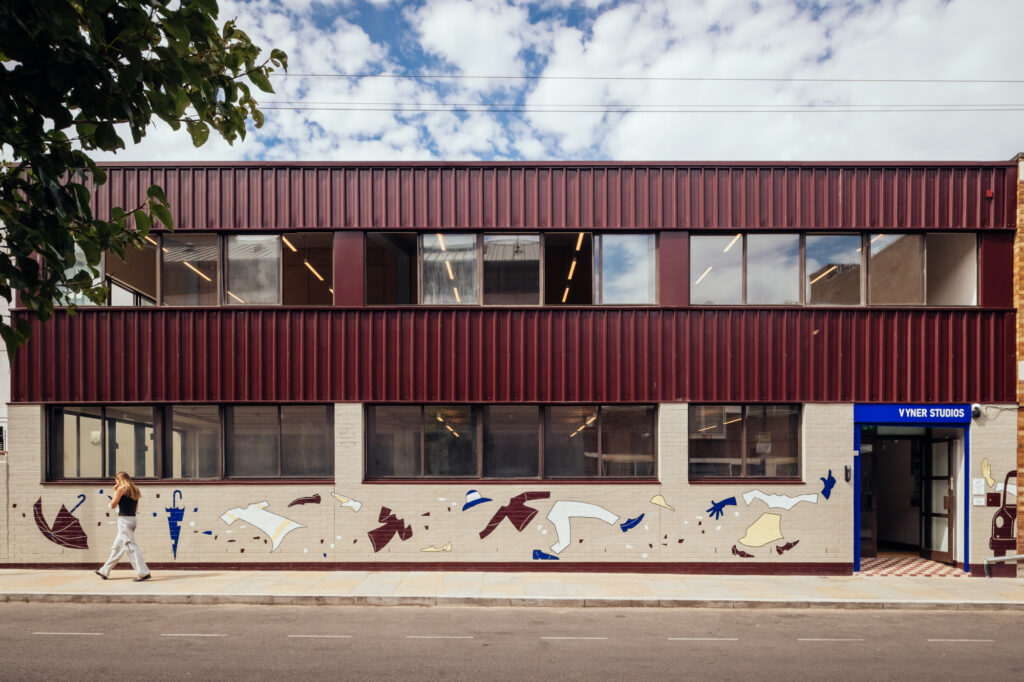 A woman walks past Vyner Studios, a two-storey building with maroon cladding, large windows, and a mural on the lower wall. The entrance reads Vyner Studios in blue, standing out against the partly cloudy sky above.