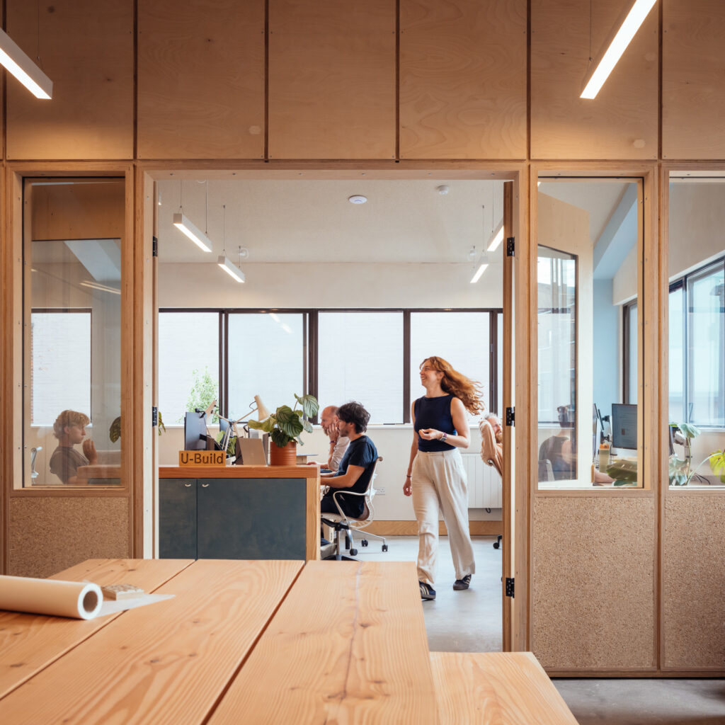 Modern office at Vyner Studios with large windows, people working at desks, and a woman walking whilst holding a phone. Some indoor plants and wooden furnishings are visible, creating a bright and collaborative workspace.