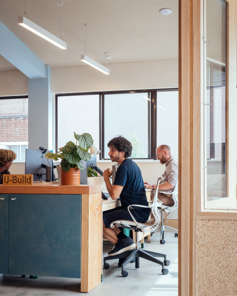 Two people work at desks in a modern office with large windows at Vyner Studios. One person faces a computer, while the other looks ahead. A potted plant sits on the counter beside them. Natural light fills the bright workspace.