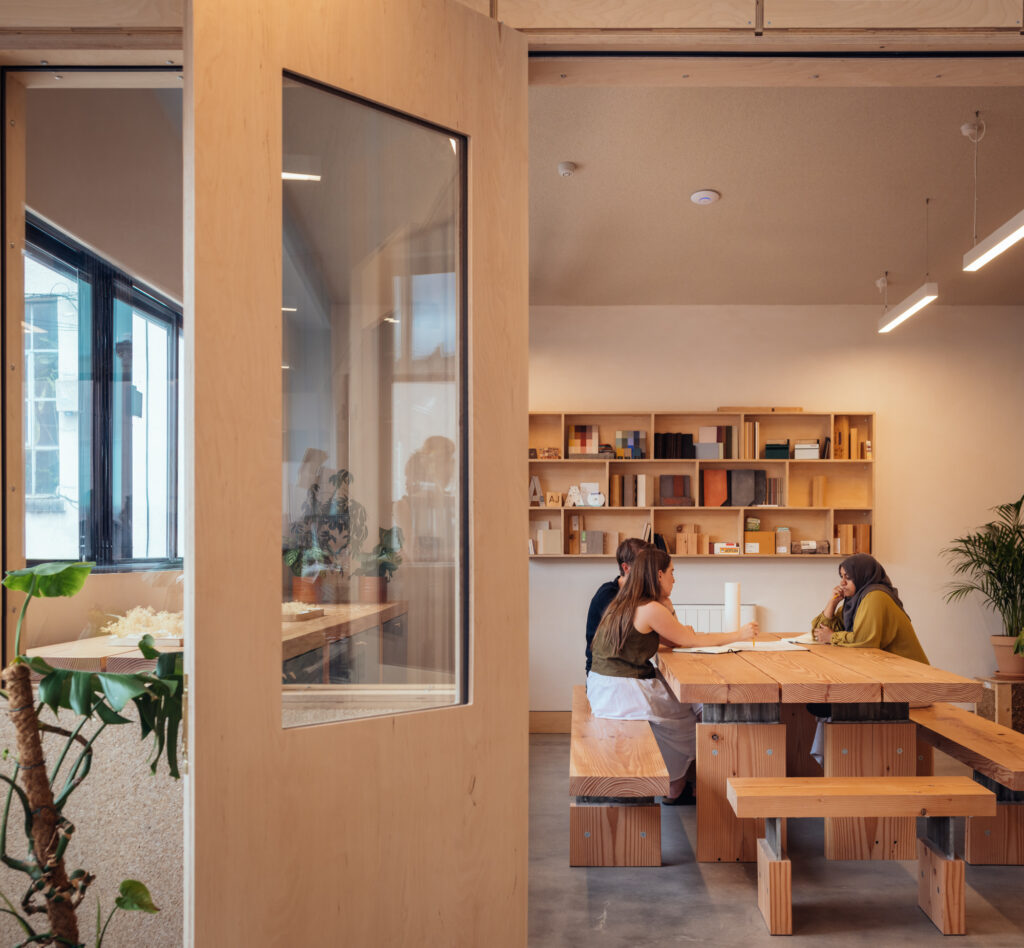 Three people sit at a wooden table in a modern, cosy room with bookshelves, plants, and large windows—reminiscent of the inviting atmosphere found at Vyner Studios—engaged in conversation. The scene is viewed through an open door.