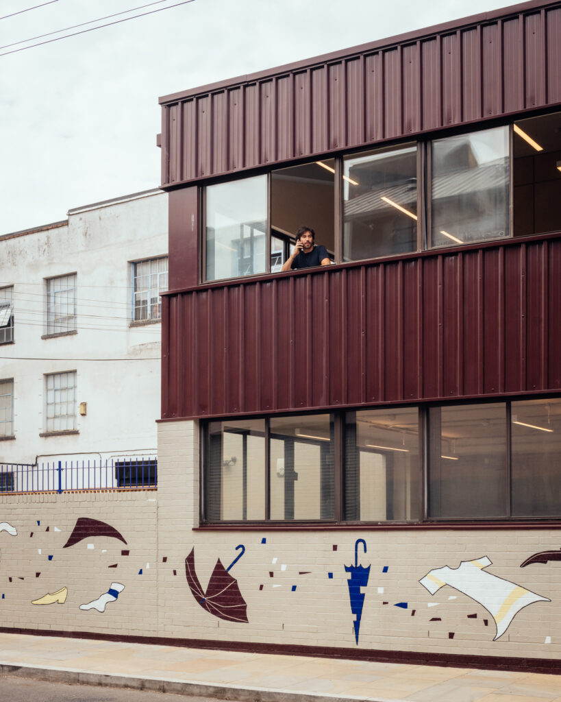 A person leans out of a first-floor window of Vyner Studios, a modern brick building with brown panels. The wall below is decorated with vibrant murals of umbrellas, hats, and flying papers.