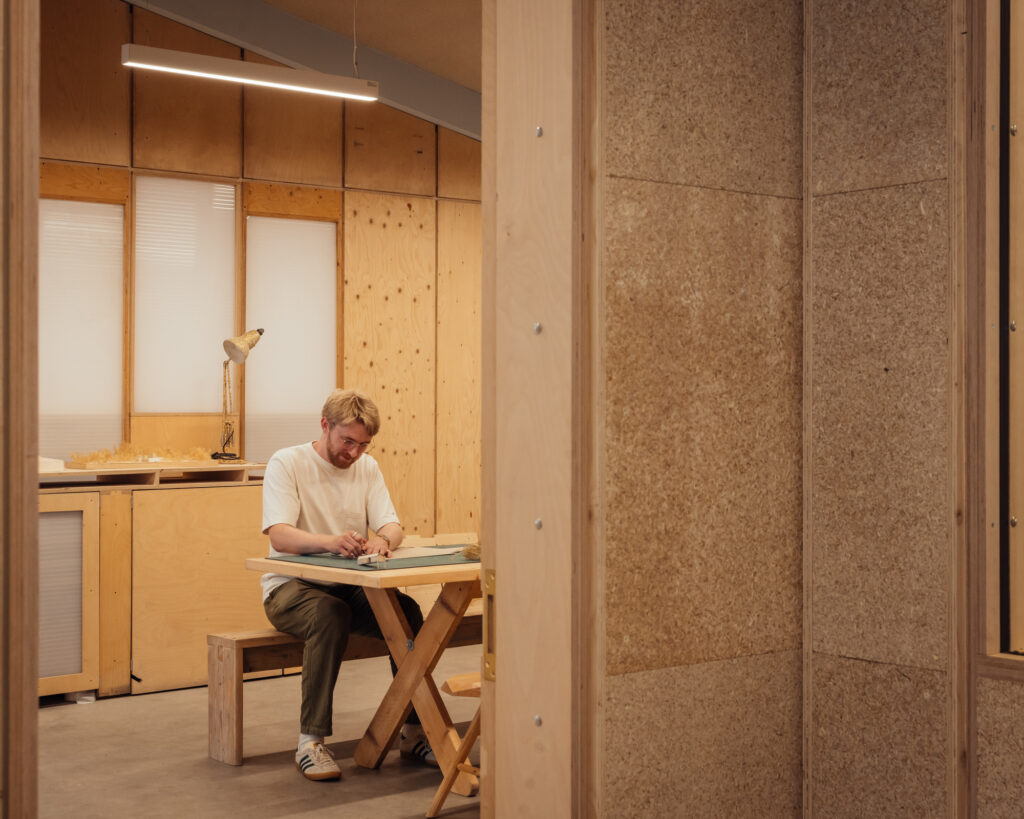 A person sits alone at a wooden table in a modern, minimalist room at Vyner Studios, working with papers. Warm lighting and wooden walls create a cosy atmosphere.
