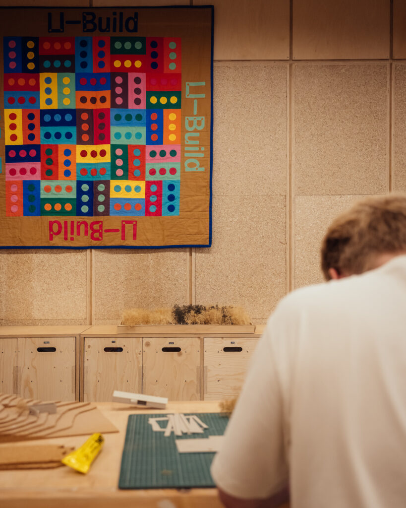 A person works at a desk with craft materials; a colourful quilt with U-Build in bold letters and geometric patterns hangs on the wall above wooden cupboards, adding creative flair to the inspiring workspace at Vyner Studios.