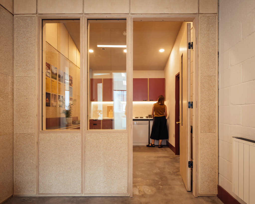 A person stands in a modern Vyner Studios kitchen with wood and red cupboards, seen through a large interior window from an adjoining room with light-coloured walls and a concrete floor.