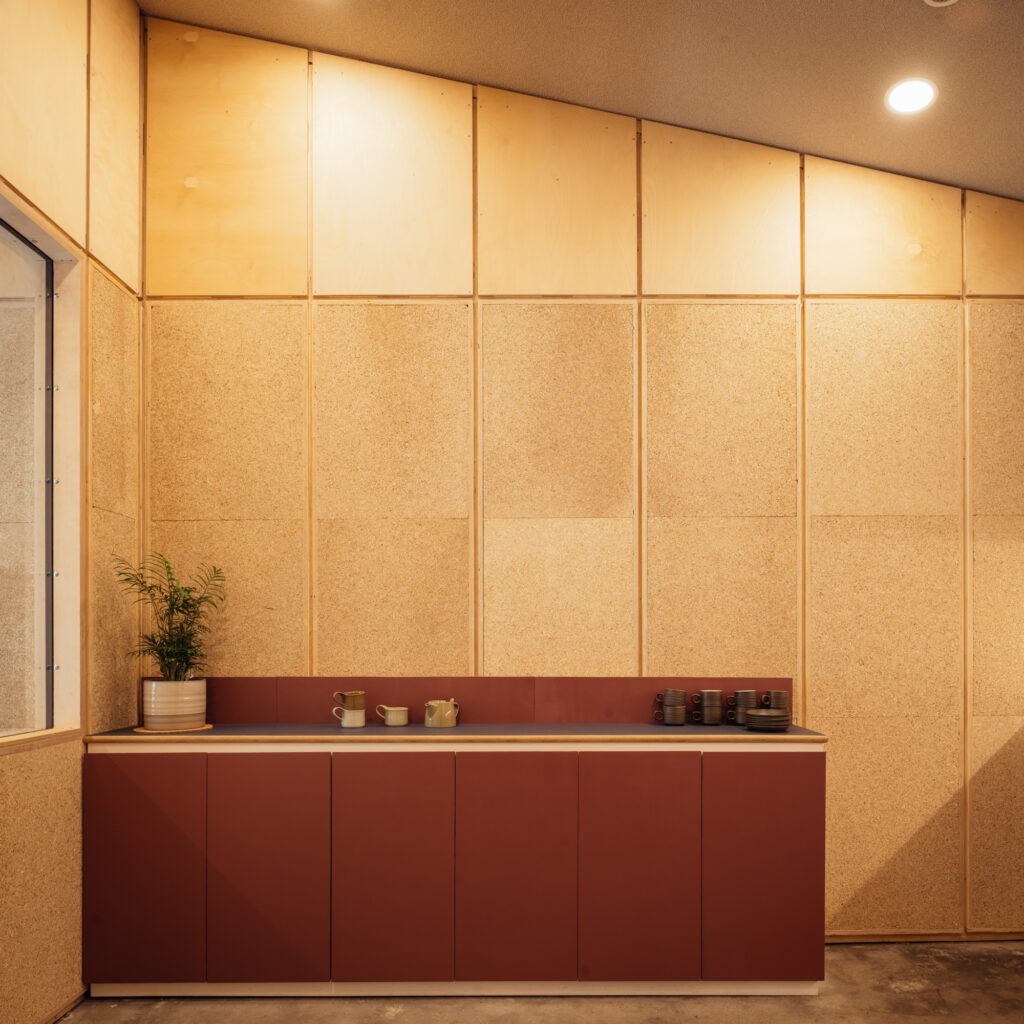 Minimalist kitchenette at Vyner Studios with red lower cupboards, beige cork walls, a plant in a striped pot, cups, and teapots on the worktop. Ceiling slopes upward at an angle. Warm indoor lighting.