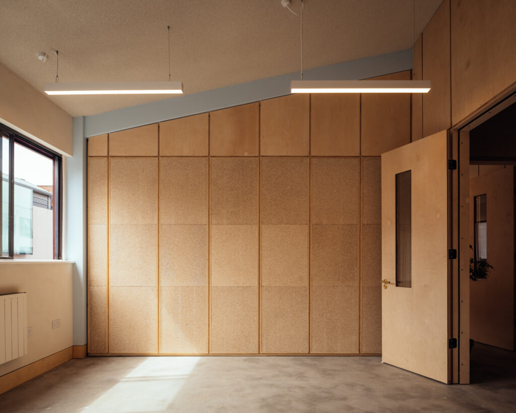 Minimalist room with wooden panelled walls, a large window on the left, two rectangular ceiling lights, and an open door revealing another room. The space is empty and has a modern design, embodying the refined aesthetic often found at Vyner Studios.