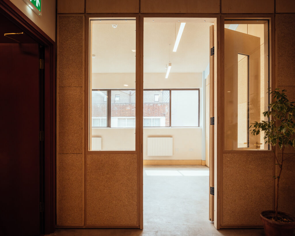 A view through glass doors into a bright, empty room at Vyner Studios, with large windows, beige walls, a potted plant, and a door partially open.