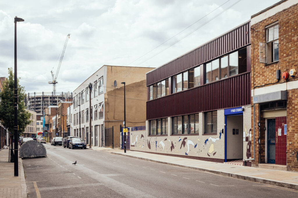 A quiet urban street with parked cars, modern buildings, and a construction crane in the distance, where Vyner Studios adds a creative touch to the scene. A mural brightens a building wall nearby, while a pigeon stands calmly in the road.