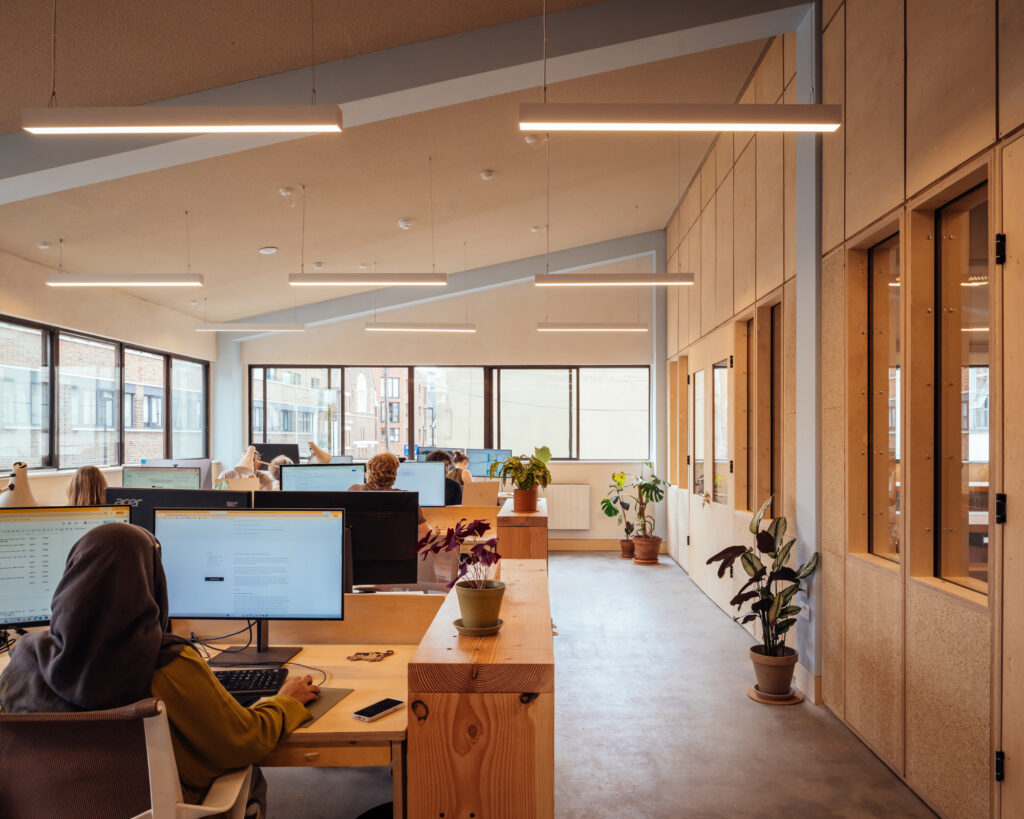 Modern open-plan office at Vyner Studios with people working at desks, large windows letting in natural light, computers, potted plants, and wooden decor elements, creating a bright and collaborative workspace.