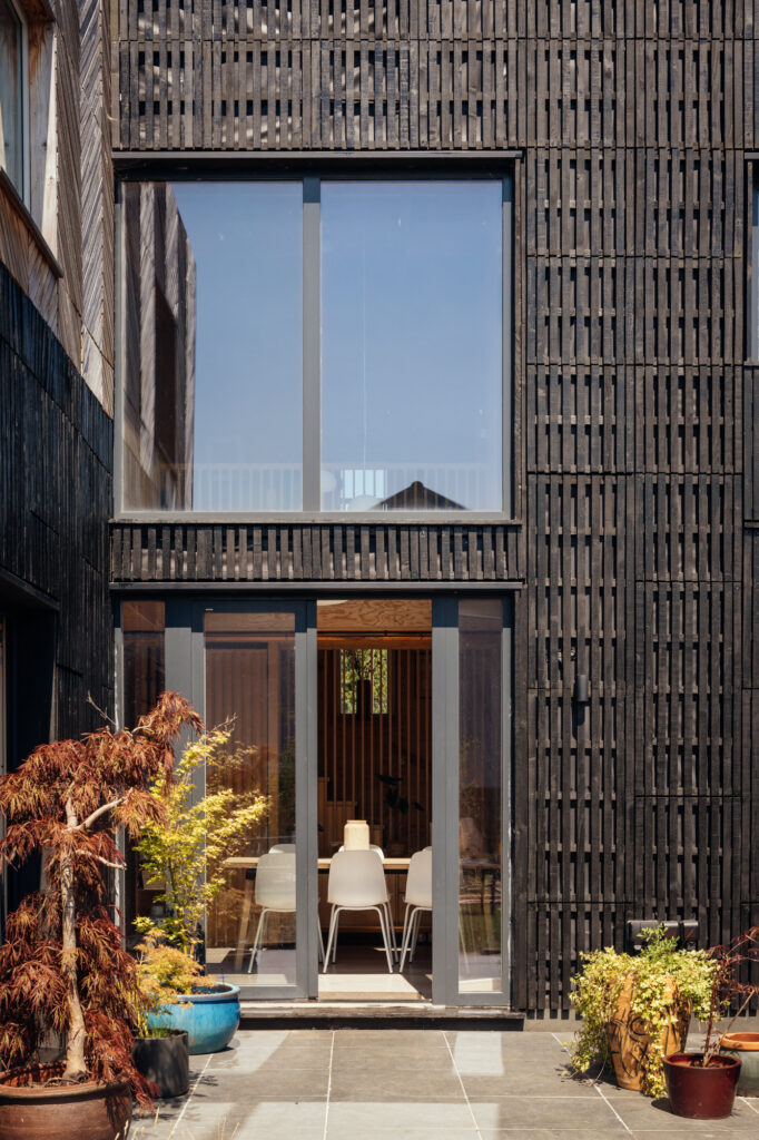 Modern house with black vertical-slat cladding and a box house extension featuring a large two-storey window and sliding glass doors opening to a stone patio with potted plants.