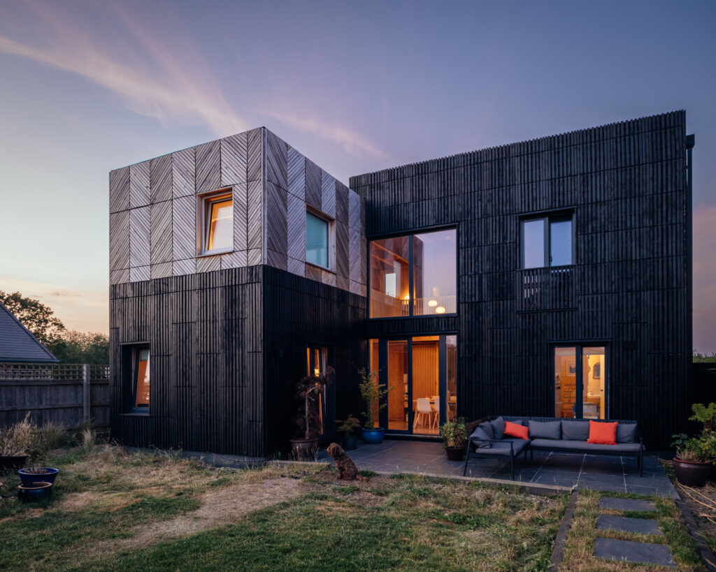 A modern two-storey home with a box extension featuring black slatted cladding and a patterned upper section, with a patio sectional sofa at dusk.