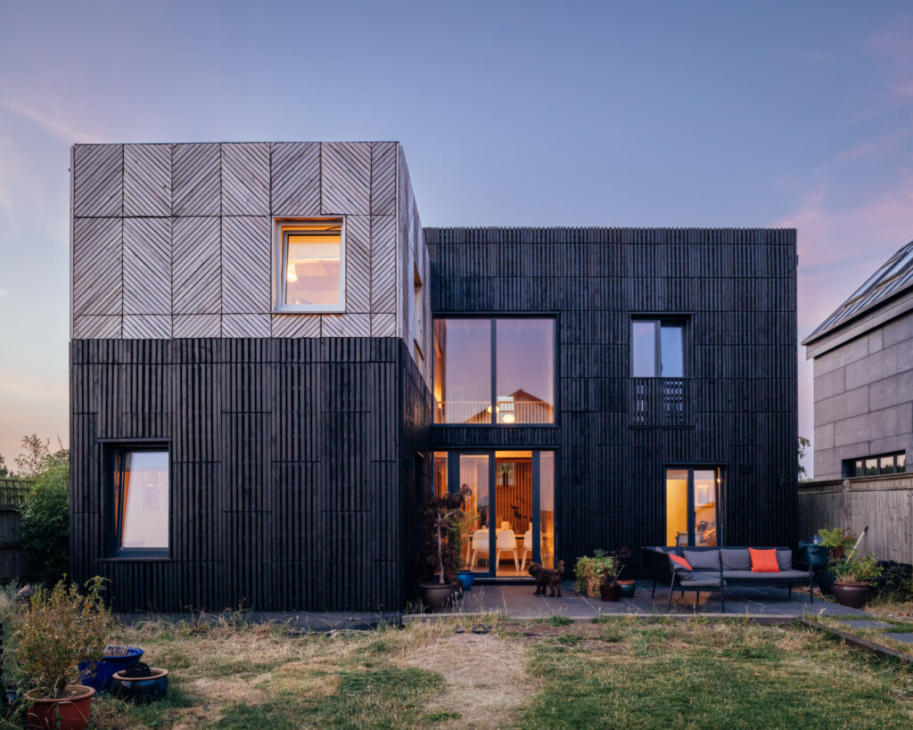 A two-storey modern house with dark vertical timber cladding and a box house extension, featuring a square upper section with chevron-patterned timber panelling, large windows, and a rear patio with outdoor seating.