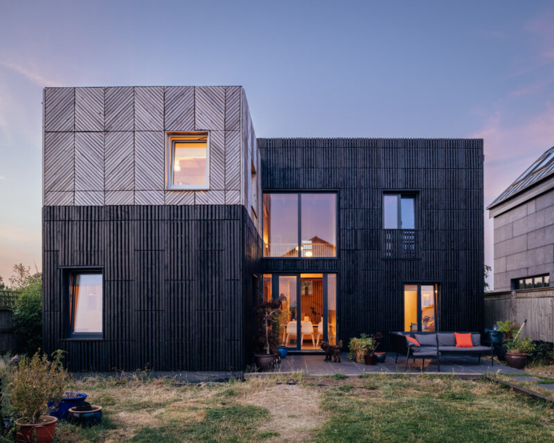 A two-storey modern house with dark vertical timber cladding and a box house extension, featuring a square upper section with chevron-patterned timber panelling, large windows, and a rear patio with outdoor seating.