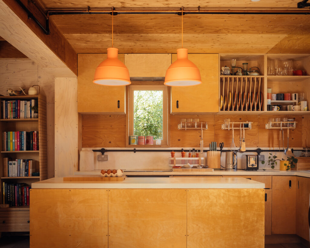 A kitchen with plywood cupboards and a central island within a box house extension, featuring two bright orange pendant lights above the worktop and a bookcase on the left.