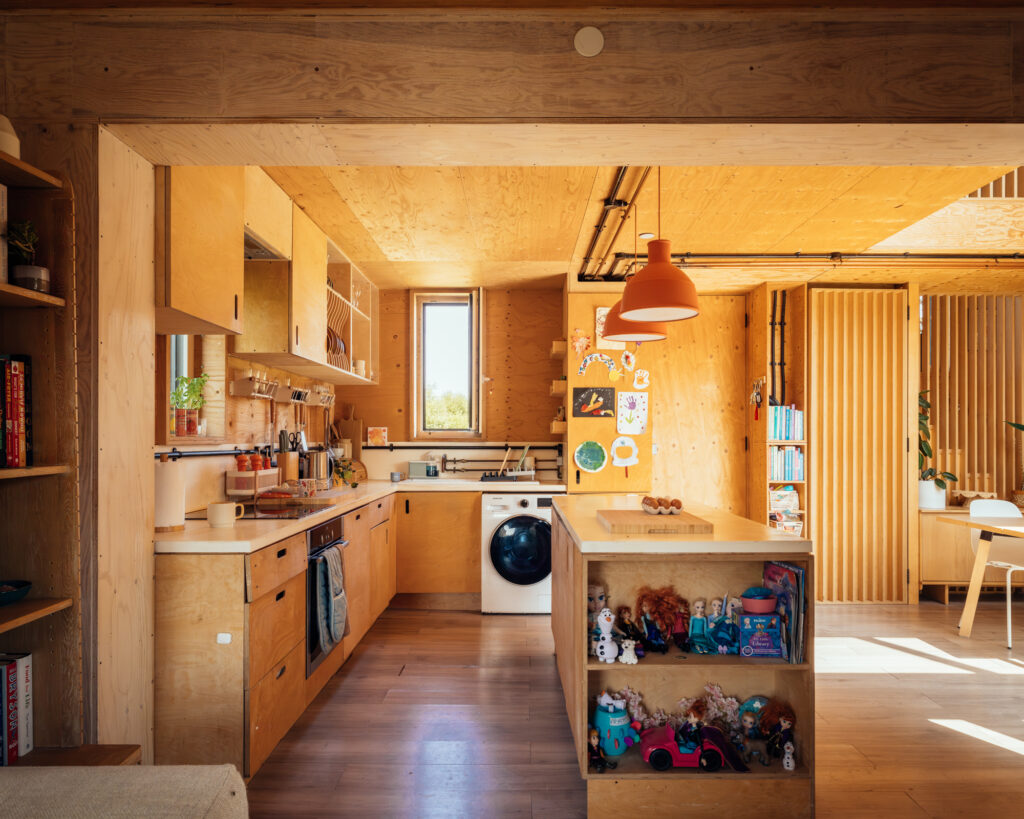 Plywood kitchen with a white worktop, built-in washing machine, and an island featuring open shelving filled with children's toys in a box house extension.