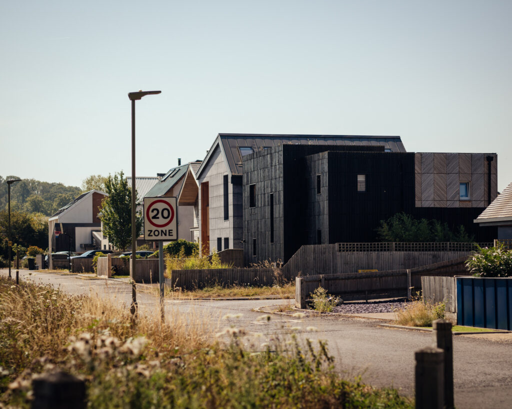 A 20 mph zone sign stands on a suburban street lined with modern homes showcasing box house extension architectural design and black and grey façades.