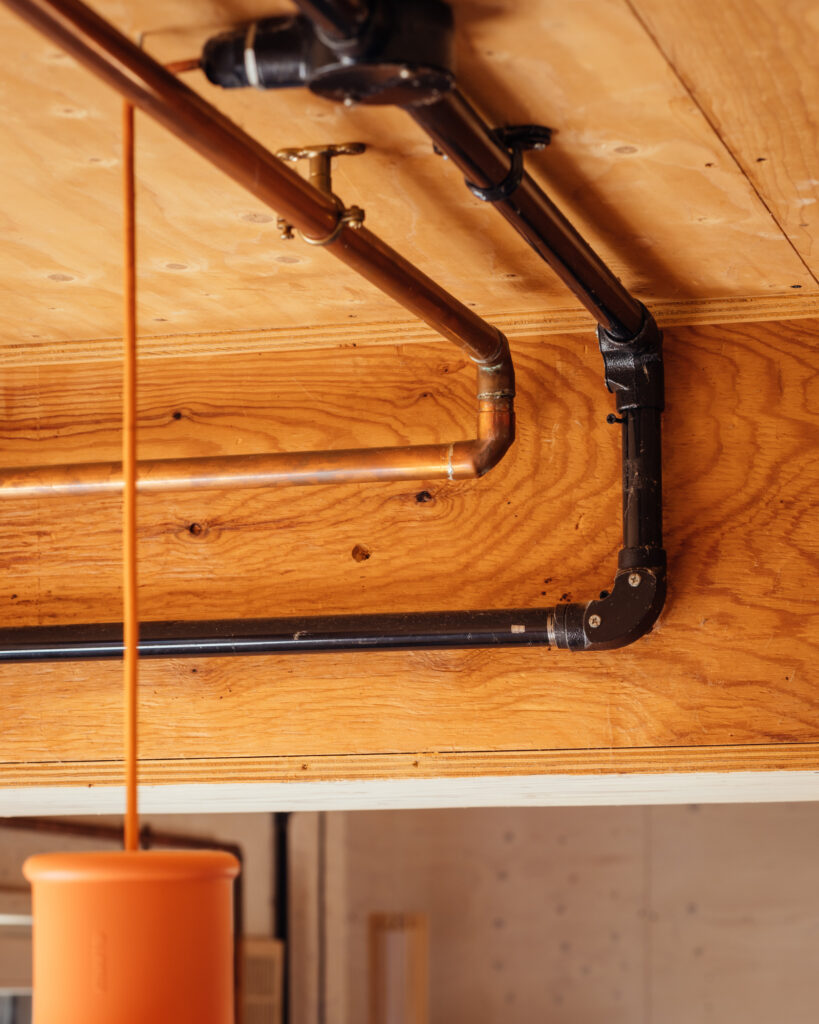 Copper and black pipes mounted against a plywood ceiling in this house extension, with a bright orange pendant light hanging in the foreground.