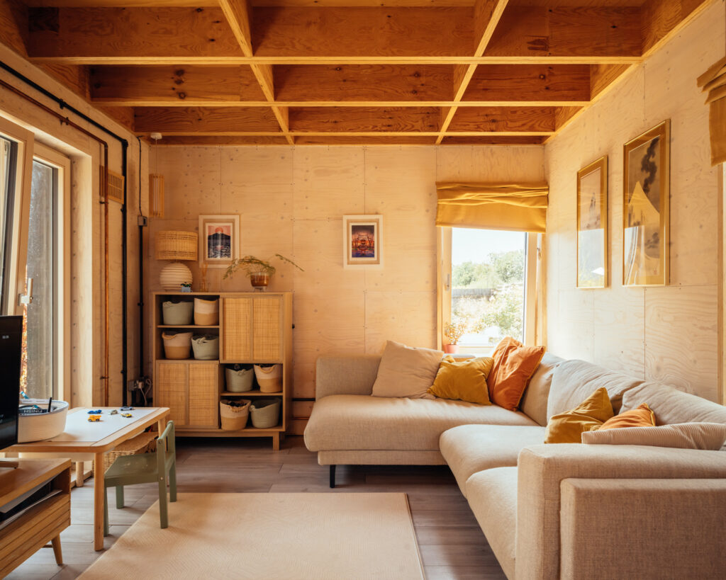 Living room within a modern home extension featuring plywood walls, a beige corner sofa with yellow and orange cushions, and a small wooden storage unit with baskets.