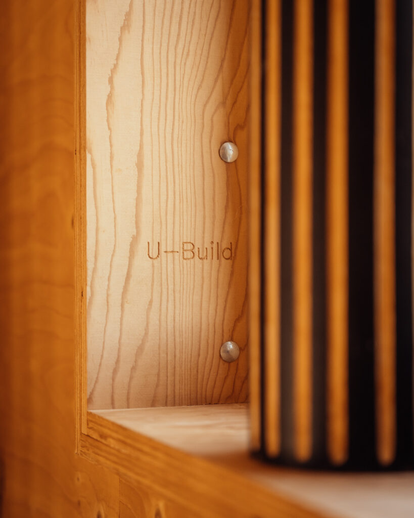 The brand name U-Build is engraved on a wooden panel of a house extension, fastened with two screws and partially obscured by a dark, vertically fluted column.