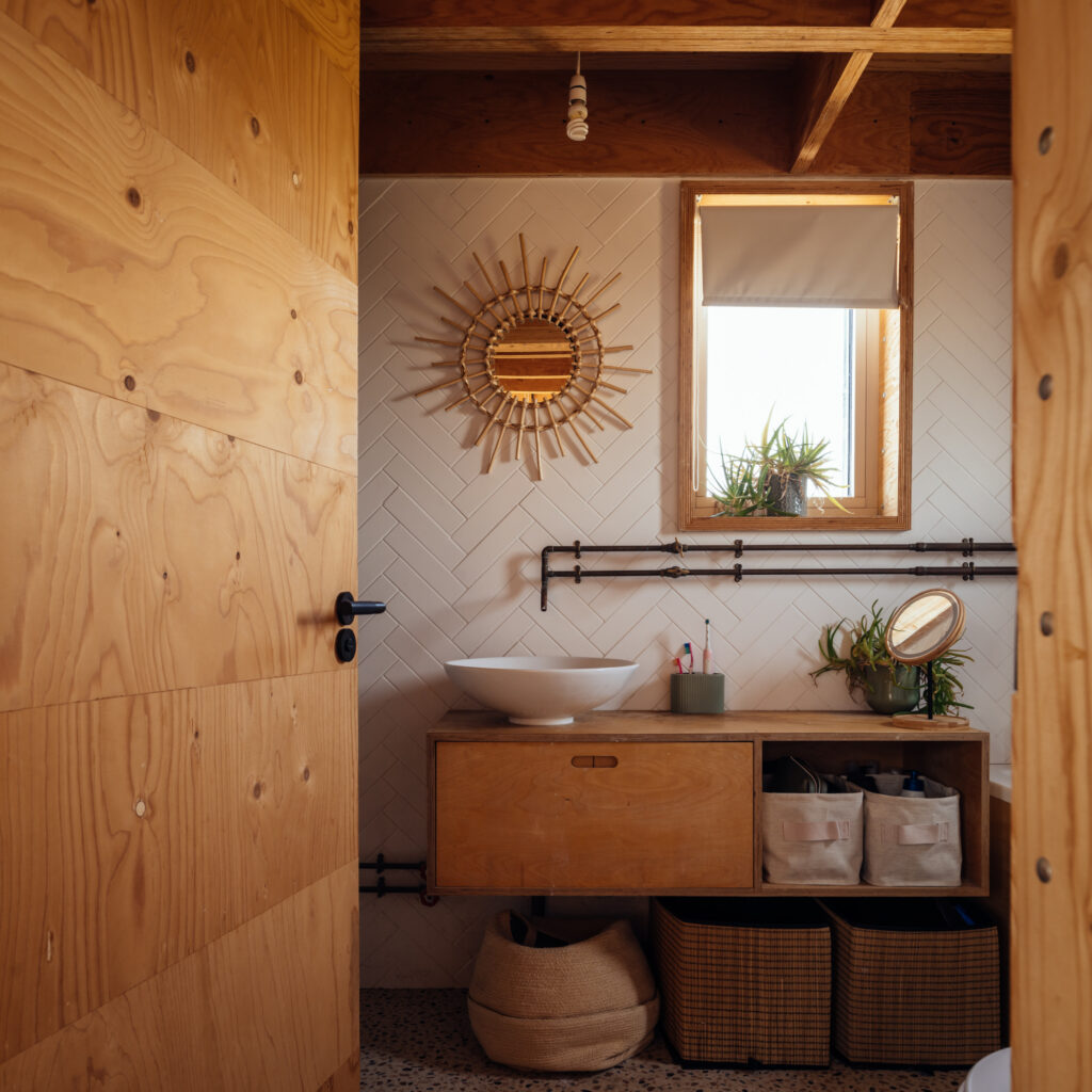 A bathroom vanity featuring a white vessel basin, a sunburst mirror on a white chevron-tiled wall, and storage baskets in a home extension.