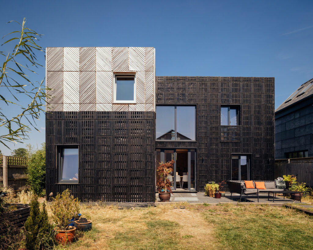 A modern two-storey home extension with a black slatted exterior and a light-coloured, chevron-patterned upper section, featuring a patio with outdoor furniture in a back garden.