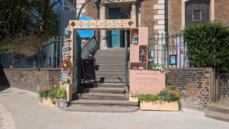 Entrance to a Sustainable Design Forum event features a wooden archway, signs, and flower boxes—an installation marking the Threshold of Change—leading up steps to a blue door with informational boards and decorations outside.