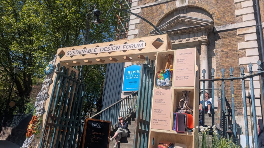 Wooden archway labelled Sustainable Design Forum marks the Threshold of Change at a brick building entrance, featuring posters on textile waste and sustainability, with colourful fabric samples in this art installation.