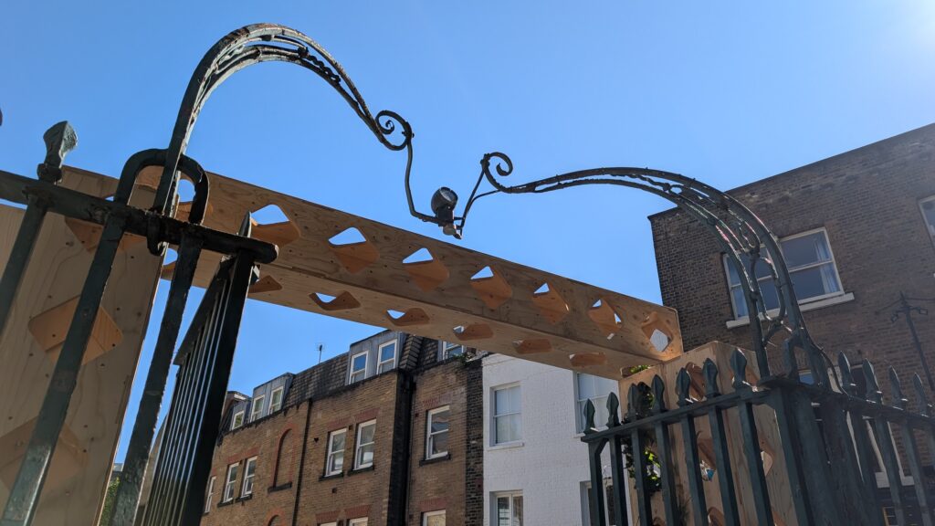 Ornate metal gate with a temporary wooden beam attached—a contemporary art installation symbolising the Threshold of Change, set against a clear blue sky and brick buildings in the background.