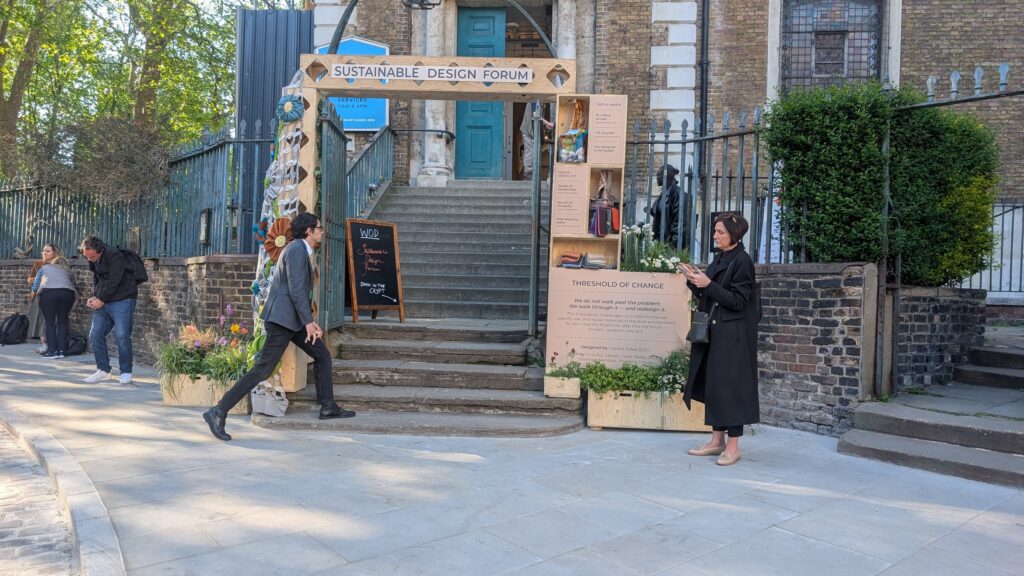People walk near an outdoor event entrance labelled Sustainable Design Forum, featuring art installations, signs, and plants—a woman in a black coat stands on the right using her mobile at this threshold of change.