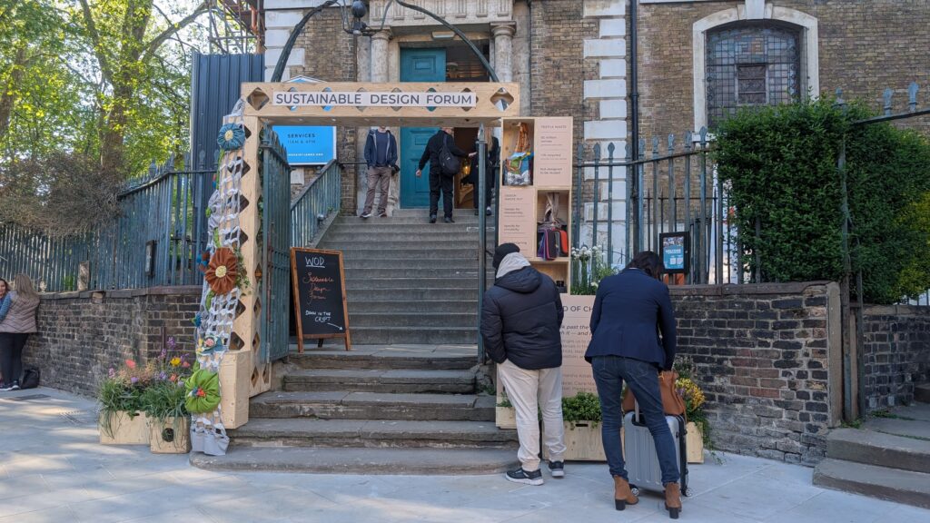 People enter a building under a wooden arch labelled Sustainable Design Forum, passing an art installation titled Threshold of Change with informational boards, plants, and decorations displayed around the entrance.