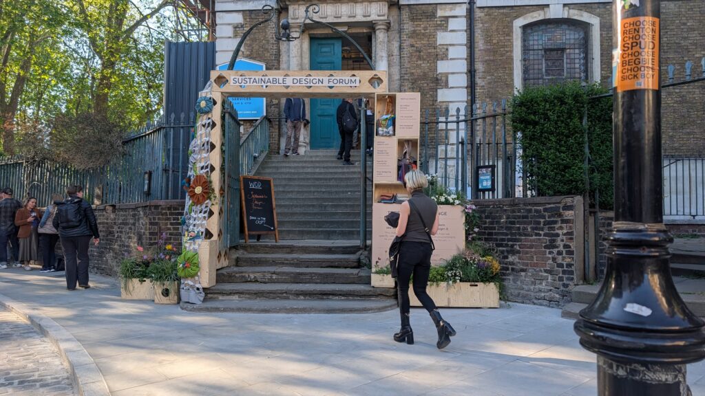 A person walks towards a decorated archway labelled Sustainable Design Forum at the entrance, where plants, information boards, and a contemporary art installation titled Threshold of Change are displayed nearby.