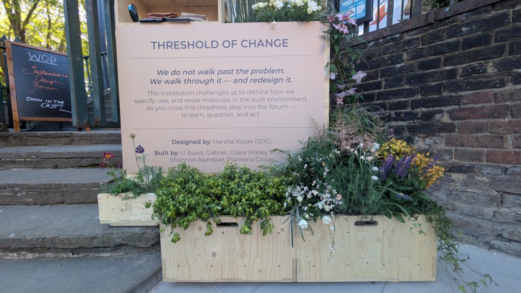A wooden planter with assorted green and flowering plants sits before the Threshold of Change installation, urging reconsideration of material use in the built environment. Brick wall background and wooden steps visible.
